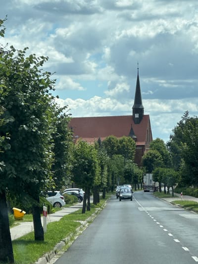 The image captures a street scene in Duszniki, Poland, featuring a prominent brick church with a dark steeple topped with a cross. The church has a red tiled roof and a clock face visible on its steeple. A line of trees with lush green foliage flanks the street on both sides. Several cars are visible on the road, including a white SUV parked on the side, a dark-colored SUV behind it, and two cars driving on the asphalt road. A large truck is also present further down the road. In the foreground, on the left side of the street, a yellow waste bin is situated on the grass next to a sidewalk. The sky is partly cloudy with a mix of white and gray clouds against a blue backdrop. The overall impression is of a suburban or small-town environment on a cloudy day.