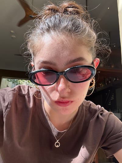 A woman with her hair in a bun wears black-framed sunglasses with red lenses. She has a nose ring and round gold earrings. A gold necklace with a circular pendant is visible against her brown t-shirt. The image is taken at an angle, showing a glimpse of the surroundings. Sunlight creates bright patches on her face and shirt.