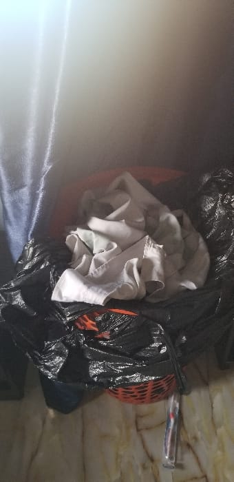 The image depicts an indoor scene focused on a collection of domestic items. The precise location within the city of Ado-Ekiti, Nigeria, is not discernible from the visual evidence presented.

In the central foreground, a pile of light-colored, possibly white or off-white, crumpled fabric, consistent with laundry, rests atop a black plastic bag. This bag appears to contain or cover an orange plastic laundry basket, parts of which are visible through perforations in the bag, revealing its grid-like pattern. To the far left, a vertical section of dark blue or navy blue fabric, consistent with a curtain or heavy drape, is present.

The background above the items is brightly illuminated and faded, suggesting an overhead light source or strong ambient light. The floor consists of light-colored, possibly beige or cream-colored, tiles with irregular, darker veining patterns, resembling natural stone like marble. On the floor, near the bottom right edge of the frame, a transparent plastic-wrapped object, consistent in shape and size with a toothbrush, is visible. No individuals are present in the scene, and no active interactions are depicted.