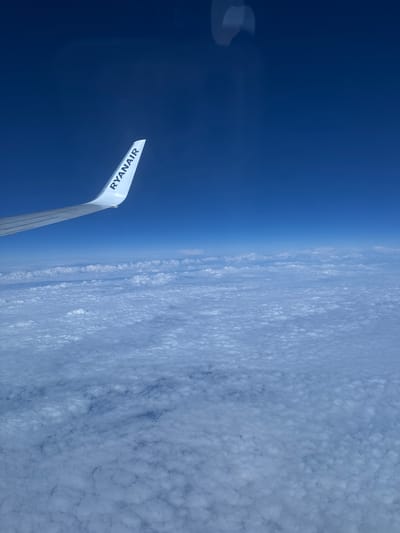 The image shows an aerial view from a Ryanair airplane. Part of the aircraft's wing, displaying the "RYANAIR" logo, is visible in the lower left of the frame. The sky is divided into two sections: on the left, a deep blue expanse, likely representing clear atmosphere at high altitude; on the right, a dense cloud cover with a textured, almost cellular appearance. The horizon line, where the blue sky meets the clouds, is roughly centered in the image. The location of Zakroczym, Poland is not apparent in the image.