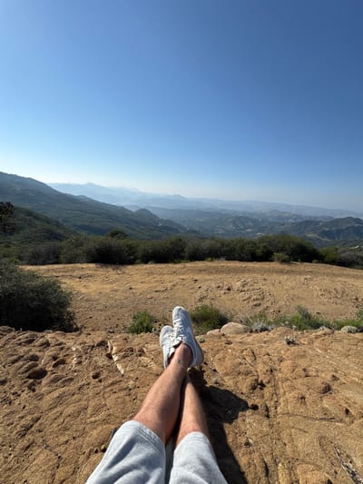 The image displays a first-person perspective from a high vantage point within the Unincorporated Santa Monica Mountains, United States of America, under a clear, bright blue sky. In the foreground, a person's bare legs, partially covered by grey athletic shorts, are extended and crossed at the ankles. The individual wears light grey sneakers with white laces. A small, dark, circular tattoo is visible on the left leg, just above the ankle. The person is resting on a textured, reddish-brown rock or dry earth surface, which exhibits cracks and unevenness, illuminated by strong direct sunlight casting distinct shadows. Beyond the immediate foreground, a wide, flat, dry, light brown dirt area is visible, interspersed with sparse patches of green and grey-green low-lying vegetation. This transitions into a dense line of dark green shrubs. The background features an expansive panorama of multiple layers of undulating mountains and hills, heavily covered in dark green foliage, receding into the distance. Evidence of distant urban development, including scattered structures and roads, is discernible within the valleys and on the slopes. A noticeable atmospheric haze obscures the farthest mountains, rendering them progressively lighter and bluer. The upper portion of the frame is a uniform, cloudless, bright blue sky.
