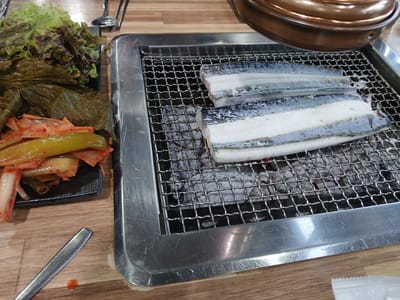 The image shows a close-up, high-angle view of a barbecue grill with three pieces of raw eel on the grate, being cooked over coals. The eel pieces are thick and white with dark blue skin. The grill is made of stainless steel and sits within a larger stainless steel tray. To the left of the grill, there is a small black plate containing a portion of kimchi, with some lettuce leaves also visible. The table surface is a light-colored wood grain. In the background, a large, copper-colored pot lid is partially visible. A metal serving utensil lies on the table to the lower left. The overall scene suggests a meal being prepared at a Korean barbecue restaurant. This scene is likely found within the city of Suwon-si, South Korea.