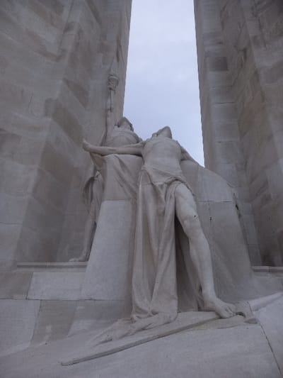 A stone sculpture features a draped figure, possibly a woman, supporting a limp figure lying across her lap. The sculpture is set against a gray sky and a building façade with rectangular stone blocks. The lower figure's leg is extended over the edge of a structure, suggesting defeat or death. The scene evokes a sense of sorrow and sacrifice.
