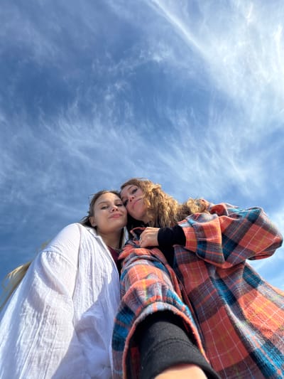 Two women are positioned in the foreground, looking down towards the camera. The background reveals a blue sky filled with wispy, white clouds. The woman closer to the camera wears a white shirt. The other woman is wearing a plaid, orange, blue, and black button-down shirt over a black, long-sleeved shirt. It is not possible to determine if the photograph was taken in Gulripshi, Abkhazia.