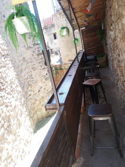 The image depicts an outdoor seating area, likely a bar or restaurant patio, in Besalú, Spain. The setting features a stone wall on the left, characteristic of the ancient architecture found in the town. On the right, a wooden slatted wall forms an enclosure. A long wooden counter runs horizontally across the foreground, with several metal bar stools with wooden seats lined up against it. Above the seating area, a wooden roof is visible, adorned with colorful triangular flags. Hanging plants add a touch of greenery to the scene, one in a white pot on the left and another in a green pot to the right. The ground is paved with concrete. The overall impression is of a quaint, rustic outdoor space, integrated into the historic environment of Besalú.