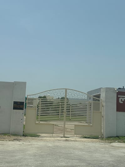 The image shows a closed, ornate, cream-colored metal gate with horizontal bars and decorative scrollwork. The gate is set between two concrete pillars. On the left pillar, a dark rectangular plaque with Hindi writing is visible. The writing appears to be "श्री वीरेन्द्र भाद" followed by some numbers that are difficult to decipher. Beyond the gate, a patch of green grass and some trees are visible, suggesting an open area or a garden. To the right of the gate, a section of a building with a dark red sign featuring a large white character, possibly "ल", is present. The sky above is clear and light blue. The scene is likely an entrance to a property in a residential or commercial area in Ajmer, India, given the script on the plaque and the architectural style. The ground in front of the gate is paved with asphalt.