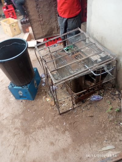 In Dokan Tofa, Nigeria, a ground-level scene captures an outdoor environment with a dirt ground surface. A person, partially visible, wears a red t-shirt and dark denim shorts. To the left, a large black plastic bin rests atop a blue plastic crate, which appears to be a beverage case. In the center of the frame, a rustic metal cage-like structure, possibly a makeshift cooler or generator enclosure, sits on the ground. This structure is covered with wire mesh on top and appears to contain some components, including a white tube. Various discarded items, including plastic wrappers and what seem to be bottle caps, are scattered around the cage and on the ground. In the background, to the left, a yellow plastic crate is visible, and further back, a tall, textured, brown rectangular object stands upright. A red plastic crate is also present in the background. The overall impression is one of an unorganized outdoor space, possibly a backyard or a service area.
