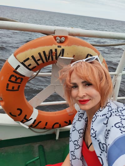 A woman with short, orangish-red hair and bright red lipstick smiles at the camera. She is wearing white sunglasses pushed up on her head and a patterned scarf draped over her shoulders. Beneath the scarf, she wears a red top. The woman is positioned in front of an orange life preserver ring that is attached to a white railing. The life preserver has black lettering, some of which is visible: "БЕРЕГ". The background is a body of water, likely a river or lake, under a cloudy sky. The scene appears to be on a boat, indicated by the railing and the presence of the life preserver. The lettering on the life preserver suggests the location is the city of Bereg in Russia.