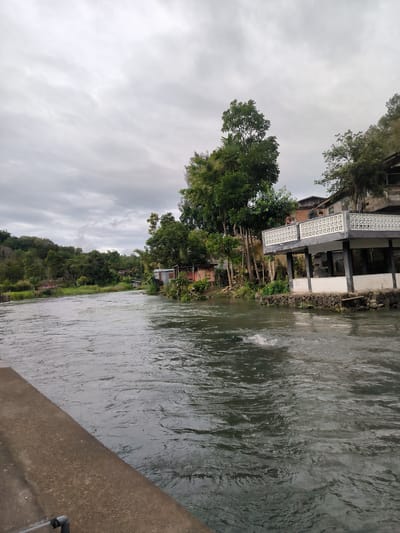 The image depicts a wide, fast-flowing river on an overcast day in Blang Bebangka, Indonesia. The river's surface is textured with ripples and a slight disturbance in the water suggests movement. On the right bank of the river, a two-story building with a white, ornate balcony stands prominently. Two individuals are visible on the balcony, looking out towards the river. Adjacent to this building are smaller structures, possibly homes or businesses, with corrugated metal roofing. Lush green trees and vegetation line the riverbanks, creating a natural, somewhat wild, environment. In the distance, rolling hills covered in dense foliage can be seen under a cloudy sky. The overall atmosphere is calm and natural, with the cloudy weather suggesting it might be late afternoon or early morning. There is no visible text in the image.