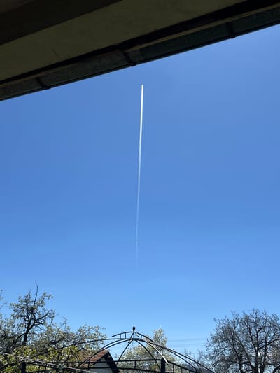 The image is a low-angle shot of the sky in Milanovo, Bulgaria. A bright blue sky dominates the frame. A contrail from a high-altitude aircraft stretches horizontally across the sky. In the lower left of the frame, a portion of a building roof is visible, with gutters. In the lower right, a decorative metal structure with flowering trees is present. The scene depicts a clear day with signs of human activity (aircraft) and both natural (trees) and built (building) environments.