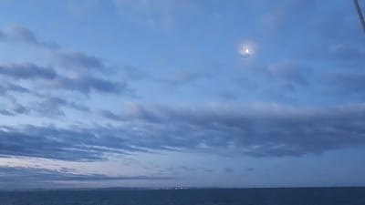 An outdoor, eye-level, wide shot captures the vast expanse of the sky above a calm body of water. The sky is a deep blue, transitioning into lighter shades of blue towards the horizon, and is partially obscured by streaks of dark gray clouds. A faint, crescent moon is visible in the upper right quadrant of the sky. The body of water, likely the sea, is a dark, deep blue and appears mostly flat, with very slight ripples. In the far distance, along the horizon, a faint line of lights suggests a distant cityscape. The overall scene evokes a twilight or early morning ambiance. No people, specific objects, or actions are discernible in the image.