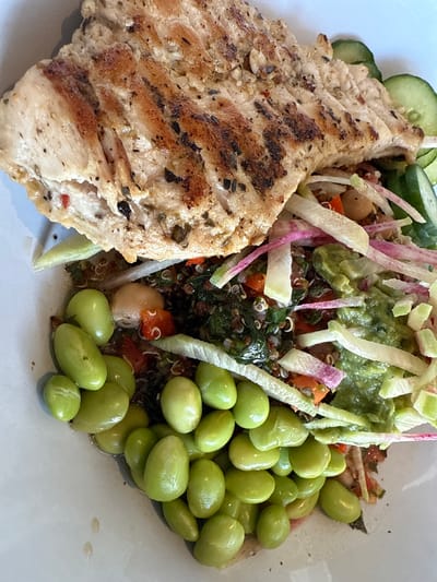 The image shows a plate of food, likely taken in Oak Brook, United States of America. A piece of grilled chicken, seasoned with herbs and spices, takes up the left side of the plate. Sliced cucumbers are positioned behind the chicken. A mixed salad dominates the upper right portion of the plate, containing ingredients such as edamame, quinoa, avocado, sliced radish, and possibly chickpeas. The arrangement suggests a prepared meal served in a restaurant or prepared food setting. The plate is white and appears to be on a table or flat surface. The lighting indicates an indoor setting with possibly natural light.