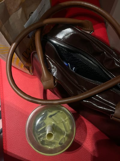 This overhead shot displays a brown patent leather handbag with a zippered opening, revealing a black interior. The bag has two rounded brown leather handles. Beside the bag sits a clear plastic cup filled with a pale green liquid and ice, with a white straw visible. The objects are placed on a textured red surface, possibly a chair or sofa. A brown paper bag with yellow and white lettering, partially obscured by the handbag, lies to the left. A white slip of paper with some faint text is also visible near the brown paper bag.

The scene appears to be indoors, with soft lighting casting gentle shadows. The overall impression is one of everyday life, with a beverage and a handbag suggesting a casual outing or a moment of rest. There is no indication of the time of day, weather, or specific emotions.

Visible text includes some faint printing on the white slip of paper and partial lettering on the brown paper bag, including what appears to be "WEST" in large yellow letters. The context of Bolesławiec, Poland, does not provide specific visual cues within the image itself.