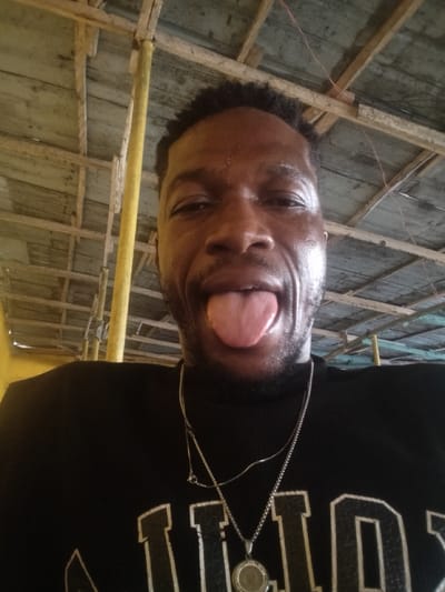 A close-up portrait shows a dark-skinned man with short, dark, curly hair and stubble or a short beard. He is looking directly at the camera with his eyes open, and his tongue is extended outwards, indicating a playful or silly expression. His face appears shiny, possibly from sweat or oil.

He is wearing a black collared shirt, which could be a t-shirt or turtleneck. Visible on the lower right side of his shirt are partial white, bold, block letters, which appear to spell out "ILLOS" or a similar ending. He has two silver-colored chain necklaces around his neck. One is a thinner, longer chain, and the other is a thicker, shorter chain with a round, decorative pendant featuring a circular pattern.

The background consists of a rustic, open ceiling or roof structure made of wooden planks and beams, suggesting an indoor or sheltered outdoor setting. The wood appears somewhat aged or weathered. On the left side of the frame, a bright yellow pole, possibly a support beam or scaffolding, is visible. Some faint red-brown wires are also visible crisscrossing in the wooden ceiling structure. The lighting is bright, illuminating the man's face. The overall mood is casual and expressive.
