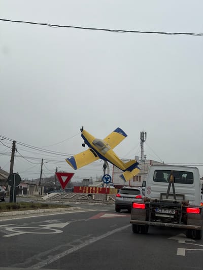 The image depicts an overcast daytime scene at a multi-lane intersection or roundabout in Tuzla, Romania, as indicated by the "AIRPORT TUZLA" sign. A prominent central feature is a yellow single-engine propeller aircraft with blue accents, mounted on a pole and positioned at an upward angle, serving as a monument. Overhead, multiple black utility lines traverse the frame.

In the foreground, a white cargo van or small truck is visible from the rear, its brake lights illuminated, indicating it is stationary or slowing. The license plate "CT 15 TJS" is clearly visible. Immediately behind this van, a silver or grey SUV is partially visible, also stopped or moving slowly, with its license plate "AG 99 SUN" discernible.

Road markings include a white left-turn arrow and red and white triangular patterns, possibly indicating a pedestrian crossing or yield area. A red inverted triangle "Yield" sign is visible to the left of the aircraft monument, alongside a blue circular "Roundabout" sign. Utility poles with numerous tangled wires are present on the left side of the road. In the background, a cellular tower or similar antenna structure is visible above residential or commercial buildings under a uniformly grey sky. No individuals are distinctly visible or identifiable.