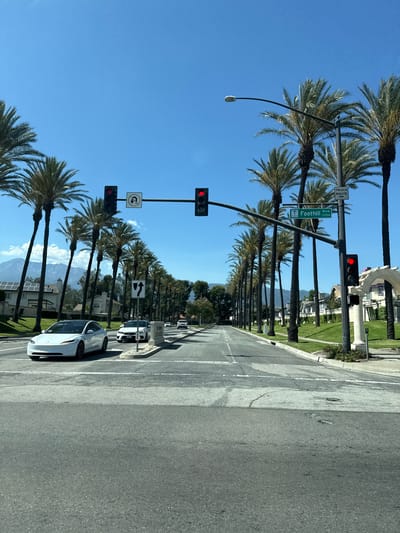The image shows a street in Fontana, California, on a clear, sunny day. The street is lined with palm trees on both sides, creating a scenic avenue. In the foreground, there are two lanes of asphalt road with white lane markings. A white Tesla is driving in the left lane. In the background, there are houses and buildings. Traffic lights are visible, with one showing a red light. A street sign indicates "Foothill Blvd 13700". A "TRUCK ROUTE" sign is also visible. Mountains can be seen in the far distance. The overall scene is typical of a suburban or semi-urban environment in Southern California.