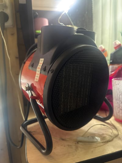 The image shows a portable electric heater on a wooden surface. The heater is red and black, with a metal grill on the front. It has a black stand. There are some labels on the side of the heater. In the background, there are some bottles and a curtain. The scene appears to be indoors, possibly in a workshop or storage area. There is no information in the image to suggest a location in the city of Разведчик, Russia.