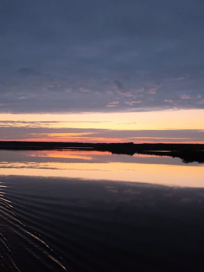 The image captures a serene landscape at dawn or dusk. The sky is a gradient of colors, transitioning from a deep blue at the top to bands of orange and pink near the horizon, with scattered clouds. Below the horizon lies a body of water, likely a lake or wide river, mirroring the colors of the sky, creating a symmetrical effect. A line of trees is present along the horizon, silhouetted against the colorful sky. In the foreground, there are ripples on the water surface, suggesting the presence of a gentle breeze or the movement of a boat. Based on the location, Даргун, Russia, the scene is likely a reflection of the surrounding natural landscape.
