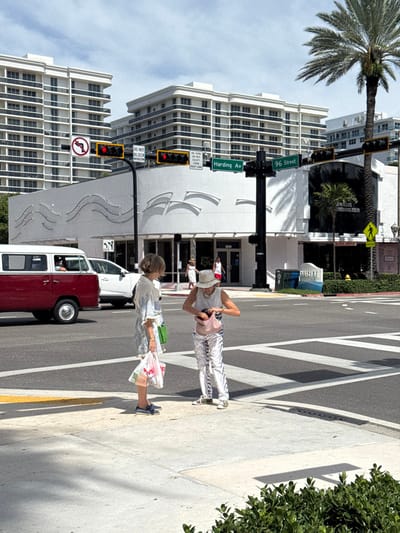 The image captures a sunlit urban street scene at the intersection of Harding Avenue and 96th Street in Bal Harbour Village, United States. In the foreground, two adult females stand on the concrete sidewalk adjacent to a pedestrian crosswalk. The woman on the left, with blonde hair, wears a light-colored patterned ensemble, carries a green shoulder bag, and holds a white plastic shopping bag with "CVS" visible. She is oriented towards the second woman. The woman on the right, distinguished by a white wide-brimmed hat, a white sleeveless top, and white and dark-striped pants, is actively rummaging through a light pink, textured handbag.
The background is dominated by multiple white, multi-story buildings, consistent with residential or hotel structures, featuring numerous balconies. A corner commercial building presents a unique white facade adorned with abstract, wave-like horizontal patterns and displays the address "1599." Adjacent to it, another commercial building has dark, reflective windows and signs for "JEWELERS & SONS.com" and "WATCH REPAIR."
The intersection is equipped with a black traffic light pole, displaying red signals for all visible directions. Green street signs indicating "Harding Av" and "96 Street" are affixed to the pole. Additional signage includes "DO NOT BLOCK INTERSECTION" and "NO TURN ON RED FROM RIGHT LANE." A vintage red Volkswagen Type 2 van and a white SUV are visible on the street, positioned as if waiting at the red light. A tall palm tree stands prominently on the right side of the frame, and green shrubs are in the immediate bottom right foreground. A white and blue decorative sign, partially reading "Bal Harbour," is visible near the intersection. The sky is partly cloudy, and strong shadows indicate a bright, high sun.