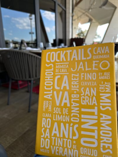 A slightly skewed close-up shot captures a yellow menu card resting on a table, likely outdoors due to the blurred background showing reflections of windows and surrounding buildings. The menu card, printed with white lettering, features a list of beverages, including words like "COCKTAILS", "CAVA", "JALEO", "FINO", "CERVEZA", "SAN", "GRÍA", "TINTO", "BLANCO", "LIMONADA" and "ANIS". The phrase "BY JOSÉ ANDRÉS" also appears on the menu card. The table is partially visible, and the blurred environment suggests an outdoor patio or dining area.
Based on the mention of "BY JOSÉ ANDRÉS" and the overall aesthetic, it is likely that the location is Jaleo at Disney Springs, in Lake Buena Vista, United States.