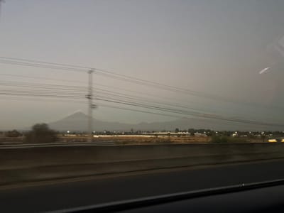 The image depicts an outdoor, hazy, twilight scene viewed from a vehicle on a highway near Santa María Zacatepec, Puebla, Mexico. In the immediate foreground, a dark vehicle interior frame borders the bottom. Above this, a dark concrete highway barrier spans horizontally. Beyond the barrier, a multi-lane asphalt road surface is visible. The midground features a flat expanse of dry, light-brown terrain with scattered low-rise buildings, possibly industrial or commercial, and sparse trees. Numerous dark overhead power lines run horizontally across the frame, supported by utility poles, including a prominent, vertically blurred pole near the center-left. Small, bright points of artificial light, some exhibiting motion streaks, are visible within the midground. The background is dominated by a large, hazy, conical mountain, identifiable as Popocatépetl, flanked by lower mountain ranges, all silhouetted against a uniform, muted gray sky. Reflections from a vehicle window are present in the upper right portion of the image. No individuals are discernible.