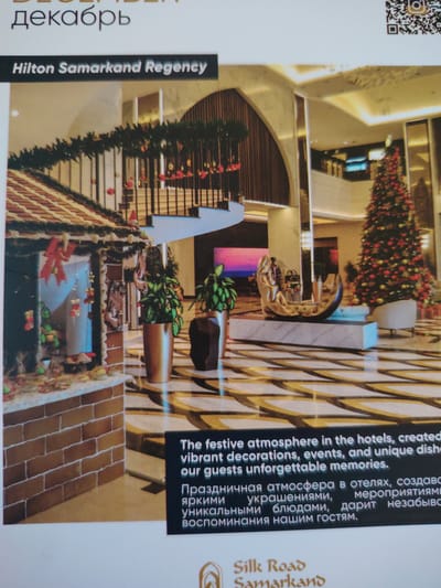 The image displays the interior of the Hilton Samarkand Regency hotel, decorated for the holiday season. A large Christmas tree adorned with red and gold ornaments stands prominently in the background. To the left, a gingerbread house facade is visible, decorated with garlands and festive trinkets. In the foreground, a stylized sculpture is placed on a marble base. The text overlays indicate that the hotel is located in Bahshitpa, Uzbekistan. The text also describes a festive atmosphere with decorations, events, and unique dishes creating unforgettable memories for guests.