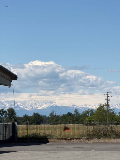 The image shows a street view in Gudo Visconti, Italy. On the right, there's a portion of a paved road, adjacent to a fence that borders a grassy field with trees. Power lines stretch across the frame, partially obscuring a view of distant mountains and a large cloud formation. The sky is blue, and a small object resembling a bird is visible in the lower left. A portion of a building or structure is also visible in the lower part of the frame.