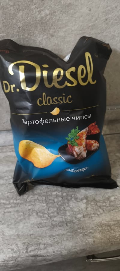 The image shows a bag of "Dr. Diesel Classic" potato chips. The bag is primarily black with gold lettering for the brand name and "classic" in a smaller font. Below that, in white lettering, it says "Картофельные чипсы," which translates to "Potato chips." A graphic of a garlic clove is positioned between the words "classic" and "Картофельные чипсы." The bottom portion of the bag features a blue gradient with an image of a potato chip and a small plate of food, possibly shrimp or prawns with herbs. The text "лобстер" (lobster) is visible on the blue part of the bag. The bag is placed on a textured, grey surface that appears to be a countertop or floor. The overall setting is an indoor environment, likely a home or store, given the plain background. The bag itself appears slightly crumpled, suggesting it has been opened or handled. There are no people visible in the image. The location in Aktanyshbash, Russia is not explicitly depicted or implied by the visual elements present.