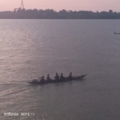 Atani, Nigeria: An eye-level, long shot reveals a group of people in a rowboat on a body of water. The rowboat is moving from left to right, with five people visible. The person in the back is operating the boat. The water ripples, suggesting movement. In the distance, a treeline sits on the horizon, with a transmission tower visible to the left. The sky is light pink and gray. There is another boat visible in the far right. The bottom left corner has the text "Infinix NOTE 11i."