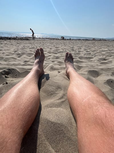 The image depicts a first-person perspective of a person relaxing on a sandy beach. The focus is on their legs and feet, which are partially buried in the sand. In the background, the sea is visible with gentle waves and a bright, sunlit sky. Two children are playing near the water's edge, one bending down and the other further in the distance. Several pieces of driftwood are scattered along the shoreline. The overall scene suggests a sunny day at the beach. The location is not identifiable as Krasnoturansk, Russia, or any other specific city based on the visual information provided.