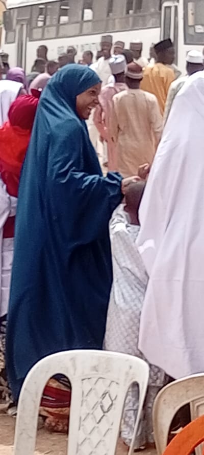 The image portrays an outdoor gathering in Rijiyar Zaki, Nigeria, under daylight conditions. In the foreground, a woman wearing a dark teal hijab and an abaya-style garment is seen from the chest up, facing right with a smiling expression. Her left hand holds the right hand or wrist of a small child positioned immediately in front of her. The child, with a bald or closely shaved head, is dressed in a white top with a subtle light grey geometric pattern, and is reaching upwards.

Behind these two primary subjects, a dense crowd of individuals is visible. The majority of the discernible adults are men dressed in traditional Nigerian attire, including various colored kaftans and caps. Several other women, some wearing hijabs, are also present within the crowd. In the background, a large, light-colored vehicle, possibly a bus or truck, is partially visible, obscured by the gathering. The ground appears to be dry and unpaved earth. The bottom foreground includes portions of at least two white plastic chairs and a hint of a third, orange-brown chair.