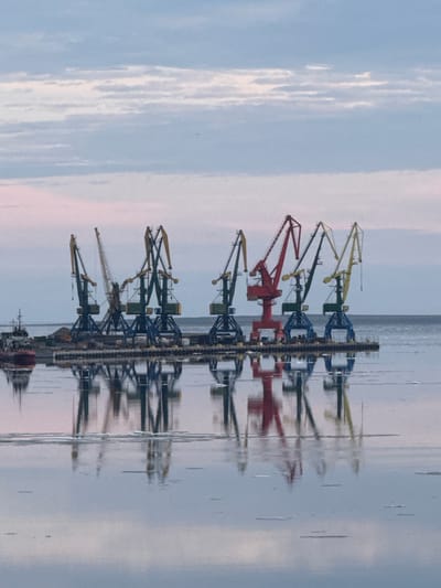 The image depicts a harbor scene in Pevek, Russia, with a row of large cranes lined up on a pier. The cranes vary in color, with some being dark green and blue, and one prominent crane being red. The sky above is overcast with pale blue and pinkish clouds, suggesting either early morning or late evening. The water in the foreground is calm and reflective, showing the inverted images of the cranes and the pier, with some ice floes visible on the surface. A red and black ship is docked on the left side of the pier. The overall atmosphere is one of industrial activity within a serene, possibly cold, environment.