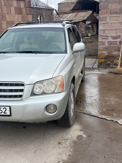 The image depicts the front and driver's side of a silver SUV, consistent with a Toyota Highlander model, parked on a concrete surface in Azatan, Armenia. The vehicle's front bumper, grille, and headlight assembly are prominent. The front bumper exhibits minor scuffs on the driver's side and appears slightly misaligned below the grille. The headlights show some hazing. A partial license plate ending in "62" is visible on the lower left of the front bumper. Roof rails are present on the vehicle's roof, and side steps are visible below the driver's door. Through the windshield, light-colored seats and a dark object hanging from the rearview mirror are discernible.
The concrete ground beneath and immediately to the right of the vehicle is wet, showing puddles and accumulated dirt/grime. A brick wall with patches of yellow-green discoloration runs along the right side of the frame, against which a natural-fiber broom leans. Beyond this wall, a dirt path leads to several dilapidated structures with weathered corrugated metal roofs and brick/stone walls. A small white object rests on the ground near these structures. In the background to the left, behind the vehicle, are other buildings constructed from red brick and stone under an overcast sky. No individuals are present, and no activity is occurring in the scene.