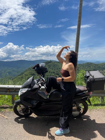 A young woman is posing on a black scooter parked on a road in Son Dien, Vietnam. She is wearing a brown bandeau top, black pants, and blue sneakers with purple laces. Her right leg is propped up on the scooter, and she is holding a pink straw in her hand, touching her forehead. The scooter has a black helmet strapped to the back and a black storage box. The background features a scenic view of lush green mountains under a blue sky with white clouds. A concrete utility pole stands to the right of the woman.