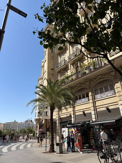 This is a street-level, eye-level, medium shot of a sunny urban scene. A light-colored building with ornate balconies and arched windows dominates the background. A palm tree stands in the middle ground, obscuring part of the building. In the foreground, a pedestrian crossing with zebra stripes is visible on the paved street. Several people are walking on the sidewalk and crossing the street. Shops with awnings are set up along the building's ground floor. To the left, a lamppost with a modern light fixture is positioned. The sky is clear and blue. The scene appears to be located in Alboraia, Spain, based on the architectural style and palm tree, which are common in Mediterranean coastal cities.