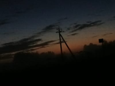 The image depicts a dusky, overcast sky above a dark, silhouetted landscape. In the mid-ground, a utility pole with wires is visible against the gradient of the sky, which transitions from deep blue at the top to orange and pink near the horizon. A small, dark rectangular sign is positioned to the right of the utility pole. The foreground is entirely black, suggesting it is obscured by darkness. The scene is devoid of any visible human presence or activity. The overall impression is one of twilight or early dawn in a rural or semi-rural setting.