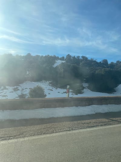 The image captures a winter scene in Ait-Oufella, Morocco, depicting a mountainous landscape under a clear blue sky with wispy clouds. The upper two-thirds of the frame show a hillside partially covered in snow, with numerous dark green trees scattered across the slopes and forming denser clumps higher up. Patches of darker ground are visible between the snow. In the mid-ground, a low, weathered stone wall extends horizontally, separating the snowy hillside from a paved road in the foreground. A distinct strip of accumulated snow rests at the base of this wall. On the snow-covered slope just behind the wall, slightly to the right of center, a vertical roadside marker with alternating red and white sections is visible. The paved road in the immediate foreground shows a coarse texture. Bright sunlight emanates from the upper left, creating a lens flare and illuminating the scene. No people, animals, or vehicles are present.