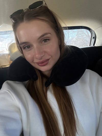 A female individual is seated within a vehicle, located in Grabovica, Montenegro. She is positioned towards the center of the frame, looking directly at the viewer with a slight smile. Her long, straight, light brown hair falls over her shoulders. Sunglasses with dark lenses and a metallic frame are perched on top of her head. She wears a white long-sleeved top or sweater and a black U-shaped travel neck pillow. The vehicle's interior features dark elements. Through the rear window, a blurred outdoor scene with tree branches against a bright sky is visible. A white, possibly textile, object is partially visible in the blurred background to the left.
