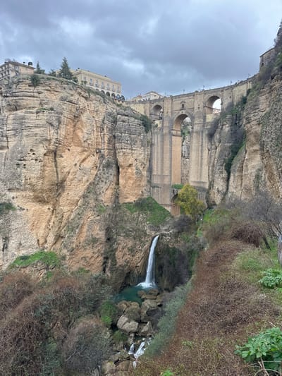 This image presents a detailed view of the El Tajo gorge in Ronda, Spain, dominated by the multi-arched Puente Nuevo bridge under an overcast sky. The massive stone bridge spans the deep chasm, featuring a large central arch and smaller ones, connecting the clifftops. Buildings of traditional architecture, including a prominent light-colored structure on the upper left, line the gorge edges. The sheer rock faces of the gorge display stratified layers in shades of tan and brown, with some pockets of green foliage. A significant waterfall cascades down the left side of the gorge, feeding into a tranquil, turquoise pool at its base, from which water continues downstream over rocks. The foreground and lower gorge slopes are densely covered with a mix of leafless shrubs and scattered evergreen plants. There are no individuals or specific human activities visible within the frame.