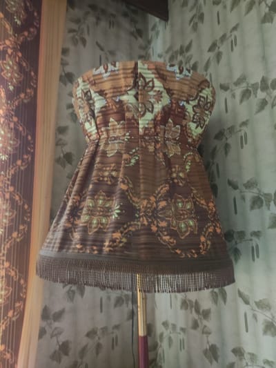 The image shows a close-up of a lampshade with a floral and striped pattern in shades of brown, orange, and green. The lampshade has fringe along its bottom edge. The lampshade is on a lamp with a metallic pole. In the background, there is a curtain with a light green leaf pattern. The lighting suggests an indoor setting. The image does not provide any information about the city of Velikiye Luki, Russia, or any specific location within it.