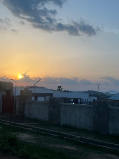 This image, captured in Rinjim Gani, Nigeria, depicts a sky transitioning from a warm, intense orange-yellow at the horizon to a lighter blue-grey overhead, indicating either sunrise or sunset. The sun is prominently visible on the left side, partially obscured by dark grey cumulus clouds, emitting distinct crepuscular rays. A substantial, light-colored concrete wall spans the mid-ground, acting as a barrier. Behind this wall, several single-story and multi-story structures, likely residential, are visible with varying rooflines and dark apertures. Two street light poles, one with a rectangular fixture, are silhouetted against the bright portion of the sky. In the immediate foreground, dark, uneven ground is present, interspersed with sparse green vegetation and concrete drainage channels running parallel to the wall. The distant background reveals a faint outline of hills or mountains on the horizon, with a utility pole and overhead lines visible on the right. No human figures or active interactions are observable within the frame.