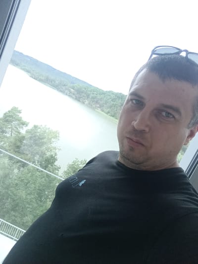 A man is taking a selfie in front of a window with a view of a lake and hills. He is wearing a black t-shirt with a blue logo that appears to say "TIDE" and has sunglasses perched on top of his head. The man has short brown hair and is looking directly at the camera with a neutral expression. The background shows a body of water surrounded by lush green trees and hills. The scene appears to be set in the city of Doksy, Czechia, known for its picturesque lake.