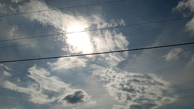 The image captures a wide-angle view of a sky filled with clouds and a bright sun. Four dark, horizontal lines, likely power lines, traverse the frame. The sun, a brilliant white orb, is partially obscured by wispy clouds on the left side of the image, casting a strong glow. Different cloud formations are visible throughout the sky, ranging from thin, scattered clouds to larger, more dense clusters. The sky is a deep blue, contrasting with the white and gray clouds. The location is identified as Kubu, Indonesia.