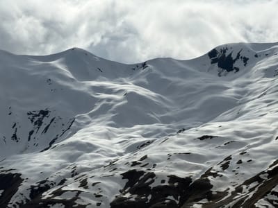 The image depicts a snow-covered mountain range under an overcast sky. Patches of dark earth are visible amidst the snow, particularly on the lower slopes. No people or signs of human activity are discernible in the frame. The location of Sioni, Georgia is not evident in this image.