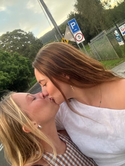 Two young women are depicted in an outdoor, head-and-shoulders profile shot, kissing with their eyes closed. The woman on the left, with blonde hair, wears a brown and white checkered top and a silver-colored teardrop earring. The woman on the right, with long brown hair, wears a white off-shoulder top and a delicate gold-colored necklace with small green beads, along with a small gold-colored earring.

The background reveals a street scene in Thirroul, Australia, likely adjacent to a school. To the right, a chain-link fence is visible, adorned with white banners, one of which prominently displays "THIRROUL." Attached to a pole are three signs: a blue rectangular "P" indicating parking, a yellow triangular warning sign depicting two children walking (a school crossing sign), and a white circular regulatory sign with a red border containing the number "5". Further in the background, green trees and foliage are present, along with a utility pole extending upwards. The sky overhead appears partly cloudy or overcast, with lighter hues suggesting twilight conditions.