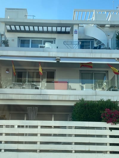 The image shows a multi-story building, likely an apartment complex, with balconies. On one of the balconies, there are two Spanish flags hanging. The building has a modern architectural style, with clean lines and large windows. There is a spiral staircase visible on one side of the building, suggesting it may be a penthouse or a design feature. In the foreground, there is a white railing. The sky is blue and clear. The location is identified as La Antigua, Spain.