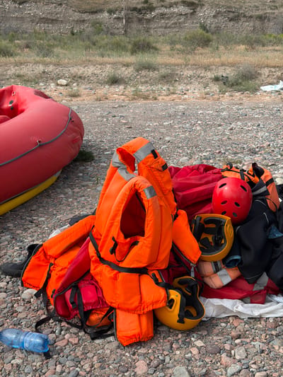 A pile of bright orange life vests and protective helmets are spread out on a rocky shore. The scene suggests preparation for a water activity such as rafting or kayaking. Among the life vests, a red helmet and two yellow helmets are visible. A large red inflatable raft is partially visible in the lower left corner of the image. The background consists of a rocky, arid landscape with sparse vegetation, typical of a mountainous or semi-arid region. A partially empty plastic water bottle lies on the rocks near the pile of gear. The lighting suggests it is daytime, likely late morning or early afternoon, with bright sunlight casting shadows. The overall impression is one of readiness for adventure in a natural outdoor setting. No people are visible in the frame, and no visible text is present on the objects.
