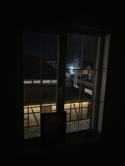 A view from a dark room through a window shows an outdoor scene at night. The window is divided into panes. Reflections are visible on the glass. Outside, there's a building with illuminated windows. The location cannot be determined from the provided image.