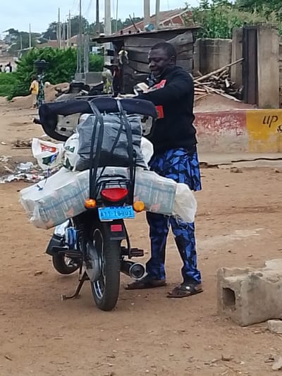 The image shows a man standing next to a motorcycle heavily laden with bags and packages. The man is wearing a black hooded sweatshirt and blue patterned trousers. He appears to be in his late 40s or 50s and is looking down at the motorcycle. The motorcycle itself is dark-colored and has a license plate that reads "AYY-783UX". Several large bags, some white and others grey, are strapped to the back of the motorcycle. Some of these bags appear to contain bottles, possibly drinks. In the background, there are several structures, including a rustic wooden building and some concrete walls. There are also piles of wood and debris. The ground is dirt and appears to be a road or a path. The sky is overcast, suggesting it might be cloudy or the early morning or late afternoon. The overall scene suggests a rural or semi-urban setting in Nigeria, likely Iseyin given the context. The man appears focused on securing his cargo or preparing for a journey.