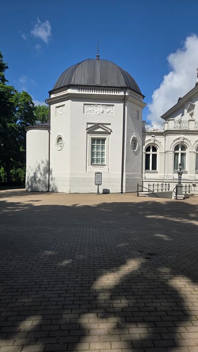 The image depicts a sunlit exterior scene, likely in Palanga, Lithuania, given the location context. The primary subjects are architectural elements of two buildings, a paved courtyard, and a clear blue sky with scattered clouds.

The foreground is dominated by a gray, domed structure with a pointed finial. This structure is attached to a larger, pale gray building with decorative relief sculptures above a large window. To the right, a white building with ornate architectural details, including arched windows and decorative railings, is visible. The courtyard is paved with light gray rectangular bricks, and dappled shadows from trees suggest it is daytime, likely late morning or early afternoon. The weather appears to be fair and sunny.

No people are visible in the frame. The scene is static and peaceful, suggesting a general view of the location rather than an event.

A small signpost is visible on the pale gray building. The text on the signpost is difficult to read clearly due to resolution and angle, but it appears to contain directional arrows and text in what might be Lithuanian.

The overall impression is one of a well-maintained historical or cultural site bathed in bright daylight.